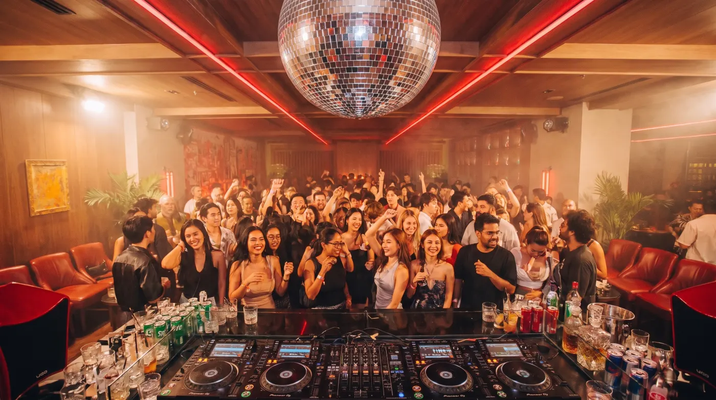 A full dance floor at Baccarat Bangkok on Sukhumvit 24, seen from the DJ booth — disco ball, red neon ceiling and a packed crowd